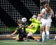 Goalkeeper diving to save a shot as another player falls near the soccer goal during an intense night game.