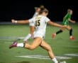 Player 15 in a white soccer uniform kicks the ball during a night game, with teammates in the background.
