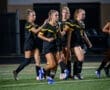 Team of female soccer players in black uniforms celebrate on the field under bright stadium lights at night.