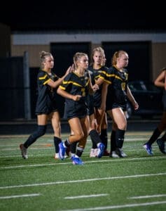 Team of female soccer players in black uniforms celebrate on the field under bright stadium lights at night.