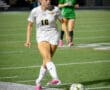 Girl in a white soccer jersey dribbles a ball on a brightly lit field during a nighttime game. Teammate in background.