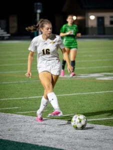 Girl in a white soccer jersey dribbles a ball on a brightly lit field during a nighttime game. Teammate in background.