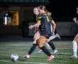 Two female soccer players in black jerseys sprinting on a field at night, gearing up for a play with a soccer ball nearby.