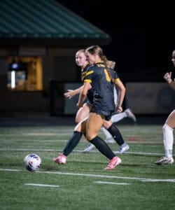 Two female soccer players in black jerseys sprinting on a field at night, gearing up for a play with a soccer ball nearby.
