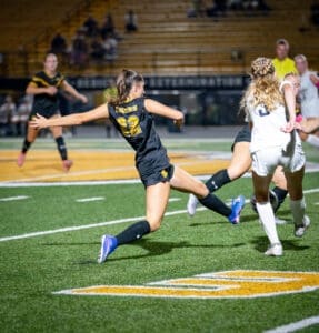 High school soccer match with player in black jersey #22 kicking the ball on the field under stadium lights.