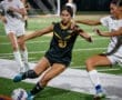 High school female soccer players compete for the ball under stadium lights on a turf field during a night match.