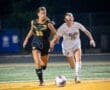 Two female soccer players compete for the ball during a night game on a brightly colored field.