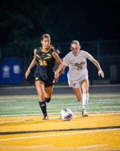 Two female soccer players compete for the ball during a night game on a brightly colored field.