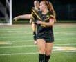 A female soccer player gestures on the field, wearing a black jersey with a pink armband during a night game.