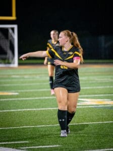 A female soccer player gestures on the field, wearing a black jersey with a pink armband during a night game.