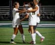 Three female soccer players celebrate a goal on the field, showing teamwork and excitement in their white uniforms.