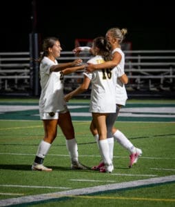 Three female soccer players celebrate a goal on the field, showing teamwork and excitement in their white uniforms.