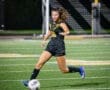 Female soccer player in black uniform dribbling a ball on a well-lit field during an evening match.
