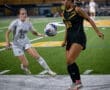Two female soccer players in a dynamic moment on the field, one dribbling a ball while the other defends actively.