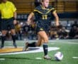 Action shot of a female soccer player in a black jersey, dribbling a ball on the field during a night game.