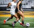 Two female soccer players competing for the ball during a night game on a turf field.
