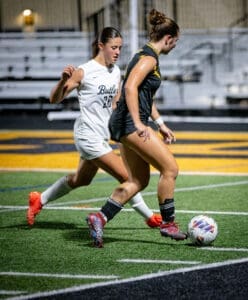 Two female soccer players competing for the ball during a night game on a turf field.