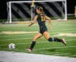 A female soccer player in a black uniform sprints toward the ball on an illuminated field during a game.