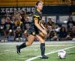 Soccer player in black uniform dribbling the ball on the field during a night game with spectators in the background.