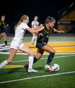 Two female soccer players compete for the ball under stadium lights during a night match on a turf field.