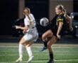 Two female soccer players competing for the ball under stadium lights during a night game on a lush green field.