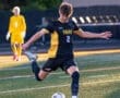 Young soccer player in a black and yellow uniform kicking a soccer ball on a field during a match. Goalkeeper in background.