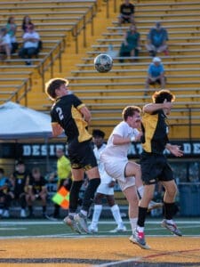 Two players jump to head a soccer ball during a match, with spectators in the background. Excitement fills the field.