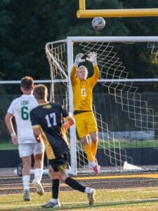 Goalkeeper leaps to save a high soccer ball during a match, with players in action around the goalpost.
