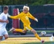 Goalkeeper in yellow jersey leaps to block a soccer ball during an outdoor match, with players in white and green uniforms nearby.