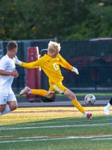 Goalkeeper in yellow jersey leaps to block a soccer ball during an outdoor match, with players in white and green uniforms nearby.