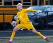A young soccer goalkeeper in yellow gear prepares to throw the ball on a field with a school bus in the background.