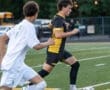 High school soccer players competing on a field, one dribbling the ball while the other defends.