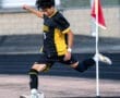 A soccer player in a black and yellow uniform takes a corner kick on a turf field.