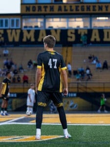 Soccer player #47 stands on the field at Newman Stadium, North Allegheny, with spectators in the background.