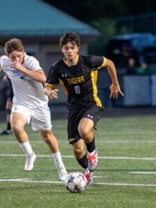 A player in a black Tigers jersey dribbles the soccer ball, pursued by an opponent in a white uniform on the field.