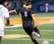 Player in black jersey kicks a soccer ball on a field, while an opponent in white follows closely behind.