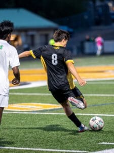 Player in black jersey kicks a soccer ball on a field, while an opponent in white follows closely behind.
