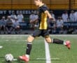Young soccer player in action, kicking a ball on a field with spectators in the background. Energetic game moment.