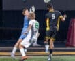 Goalkeeper leaps to save a shot while players collide on the soccer field during an intense night match.