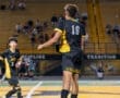 Player in black and yellow jersey jumps to head a soccer ball under stadium lights during a lively game.