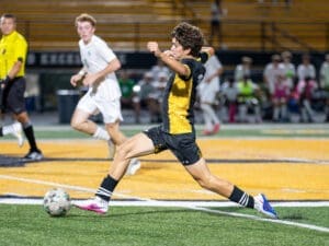A player in a black and yellow soccer jersey strikes the ball on the field during an intense game night.