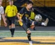 High school soccer player in black jersey strikes a ball on the field with the referee in the background.