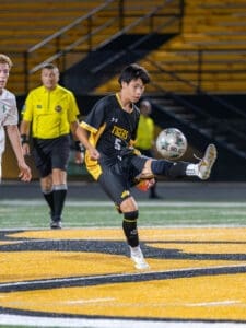 High school soccer player in black jersey strikes a ball on the field with the referee in the background.