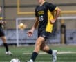 Young soccer player in black and gold uniform dribbles a ball on a field during a match, showcasing athleticism and focus.