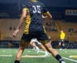 A young soccer player in a black and yellow jersey prepares to kick a ball on the field during an evening match.
