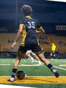 A young soccer player in a black and yellow jersey prepares to kick a ball on the field during an evening match.