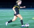 A female soccer player in a black jersey dribbles a ball on a lit field during a night game.
