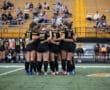 A group of female soccer players in black uniforms huddle together, showing teamwork and unity before a game.