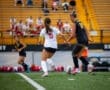 Two female soccer players compete for the ball on a field, with spectators in the background watching the match.