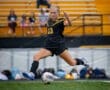 Youth soccer player in black uniform kicks a soccer ball on the field, with spectators in the background.